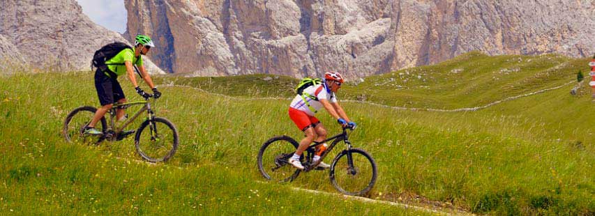 Dos ciclistas practicando deporte en bicis negras bajando por la montaña. El paisaje es verde y rocoso en el fondo.