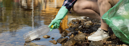 Persona con guantes de látex recogiendo residuos (botella de plástico) del río para introducirlos en una bolsa de basura que sostiene.