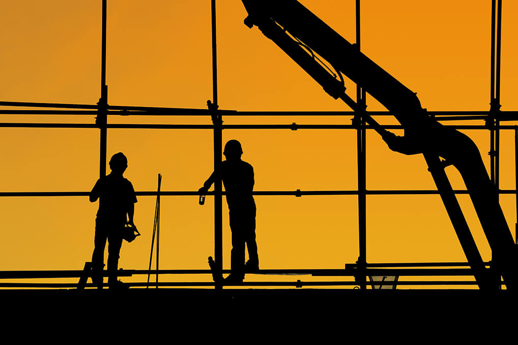 Silueta de dos trabajdores de la construcción verde encima de un andamio en el atardecer. De fondo se ve el cielo naranja.